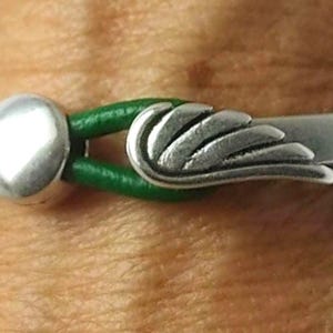 May include: Close-up of a bracelet featuring a silver wing charm and a silver round bead. The bracelet has a green leather band. The wing charm is detailed and the bracelet is set against a skin tone background.