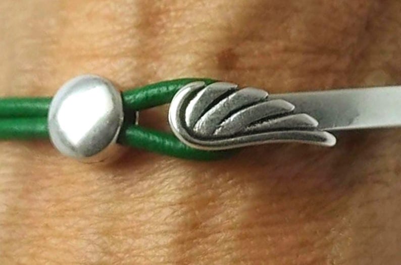 May include: Close-up of a bracelet featuring a silver wing charm and a silver round bead. The bracelet has a green leather band. The wing charm is detailed and the bracelet is set against a skin tone background.