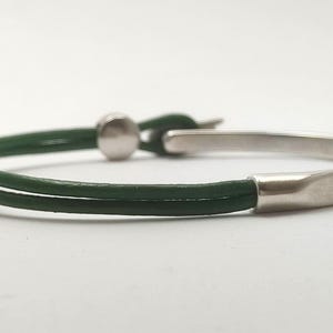 May include: A minimalist bracelet featuring a dark green leather band and a silver-colored metal clasp. The bracelet has a simple, elegant design, suitable for everyday wear. The band is approximately 0.25 inches wide.