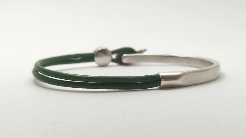 May include: A minimalist bracelet featuring a dark green leather band and a silver-colored metal clasp. The bracelet has a simple, elegant design, suitable for everyday wear. The band is approximately 0.25 inches wide.