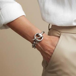 Op de afbeelding: Een zilverkleurige metalen kettingarmband met een opvallende knevelsluiting. De armband bestaat uit een dikke, gepolijste cirkelvormige ring en een bijpassende horizontale staaf die door de ring schuift om de kettingschakels aan elkaar te bevestigen.