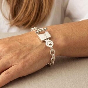 Könnte beinhalten: Ein silbernes Armband mit einem Vorhängeschloss-Design und einem Knebelverschluss. Das Armband hat einen Ketten-Look und einen kleinen runden Anhänger. Das Vorhängeschloss hat die Nummer "24" eingraviert.