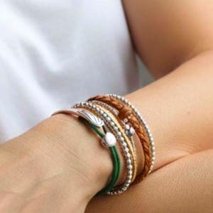 May include: A collection of layered bracelets in various styles and colors. The bracelets include silver beaded, braided leather, and green cord designs. Some bracelets have decorative charms and a metallic finish.