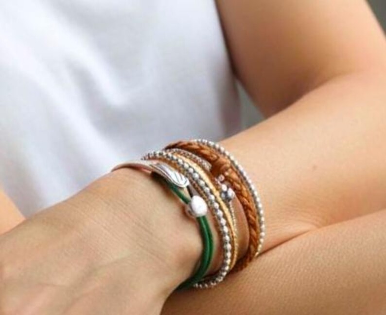 May include: A collection of layered bracelets in various styles and colors. The bracelets include silver beaded, braided leather, and green cord designs. Some bracelets have decorative charms and a metallic finish.