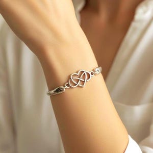 Op de afbeelding: Een zilverkleurige armband met een centrale oneindigheidshart-bedel. De bedel bestaat uit twee in elkaar verstrengelde hartvormen die een oneindigheidslus vormen. De armband is opgebouwd uit gladde, gepolijste metalen schakels die verbonden zijn met het centrale hartstuk, wat zorgt voor een doorlopende, strakke en reflecterende uitstraling om de pols.