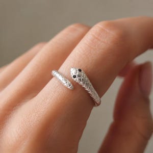 May include: A silver-toned wrap ring shaped like a snake, featuring a textured scale pattern along the band and two small, dark, circular accents representing the eyes on the head of the snake. The ring is open-ended, allowing for adjustment.