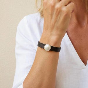 Può includere: Un bracciale in pelle testurizzata marrone scuro con una singola perla bianca rotonda incastonata in una montatura in metallo color argento al centro. Il cinturino presenta un sottile motivo in rilievo e si chiude con un fermaglio metallico.