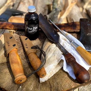 May include: A collection of outdoor tools and accessories, including two knives in tan sheaths, an axe, a bottle of Pine Fir product, and a sharpening tool. The items are arranged on a wooden surface, suggesting a rustic or outdoorsy theme.