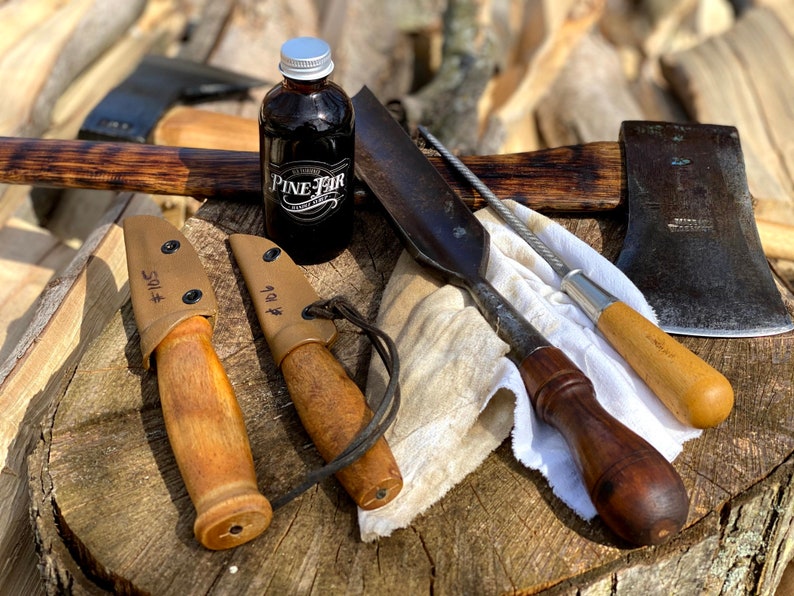 May include: A collection of outdoor tools and accessories, including two knives in tan sheaths, an axe, a bottle of Pine Fir product, and a sharpening tool. The items are arranged on a wooden surface, suggesting a rustic or outdoorsy theme.