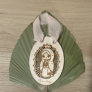 May include: A wooden oval ornament with a laser-cut design of the Virgin Mary. The ornament is decorated with a crown, stars, and a floral pattern. The name "Penelope" is written below the image.