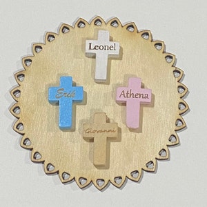 May include: Four wooden crosses with names engraved on them. The crosses are painted in different colors: white, blue, pink, and brown. The crosses are arranged on a wooden circle with a scalloped edge.