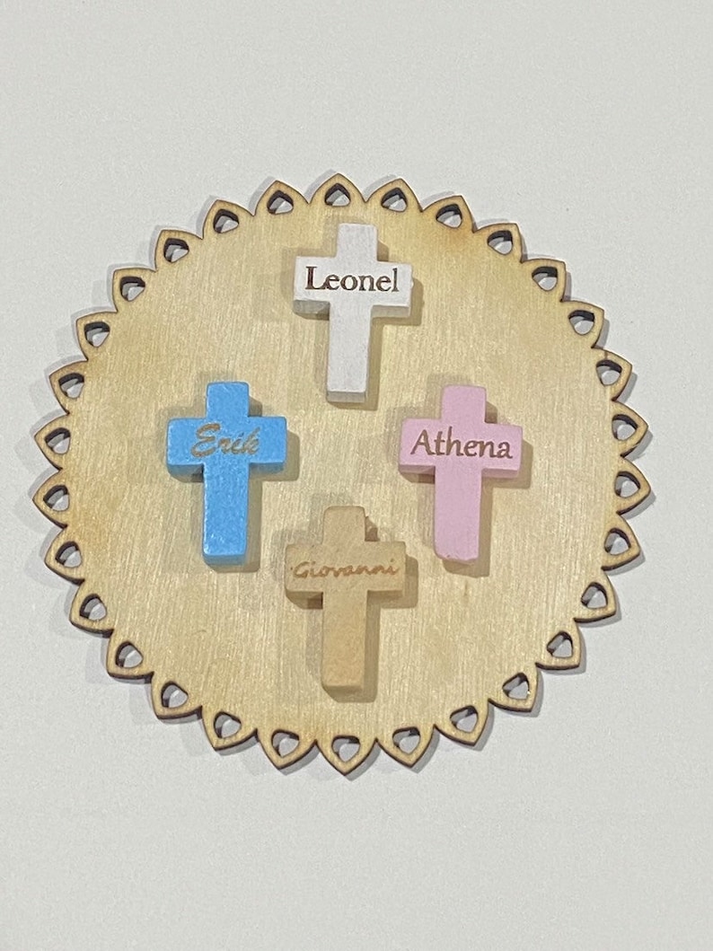 May include: Four wooden crosses with names engraved on them. The crosses are painted in different colors: white, blue, pink, and brown. The crosses are arranged on a wooden circle with a scalloped edge.
