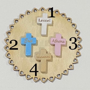 May include: A wooden circle with a scalloped edge and four wooden crosses with names on them. The names are Leonel, Erik, Athena, and Giovanni. The crosses are numbered 1 through 4.