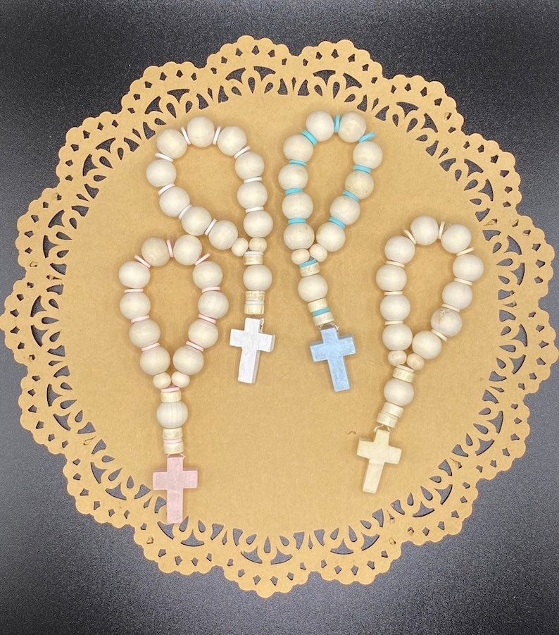 May include: Four wooden bead rosaries with different colored crosses. The rosaries are white with a light brown wood tone. The crosses are pink, blue, and light blue.