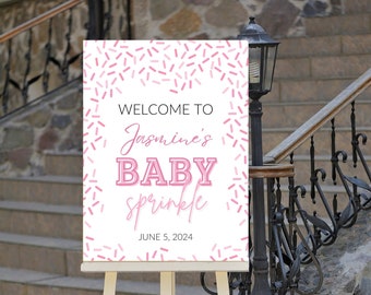 Op de afbeelding: Een wit bord met roze sprinkles op een houten ezel. Het bord zegt "Welkom bij Jasmine's Baby Sprinkle 5 juni 2024".