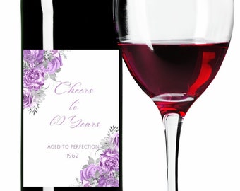 Può includere: Una bottiglia di vino con un'etichetta che dice "Cheers to 60 Years Aged to Perfection 1962" con fiori viola e dettagli argentati. Un bicchiere di vino rosso è accanto alla bottiglia.