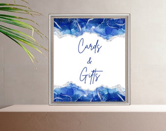 May include: A white sign with blue and silver abstract art border. The sign says "Cards & Gifts" in blue cursive text.