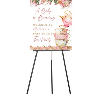 May include: A baby shower sign on a black easel. The sign has a floral design at the top and bottom. The text reads "A Baby is Brewing" and "Welcome to Melissa's Baby Shower Tea Party". The sign features a teapot and teacups.