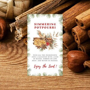 Op de afbeelding: Een witte kaart met groene dennentakken rond de rand. De kaart heeft de tekst "Simmering Potpourri" in rode letters. De kaart heeft ook een aquarel-illustratie van kaneelstokjes, sinaasappelschijfjes, steranijs en kruidnagel. De tekst onder de illustratie luidt: "Alle ingrediënten in een pan doen met 2 kopjes water. Laat op laag vuur sudderen. Voeg indien nodig water toe. Geniet van de geur!"