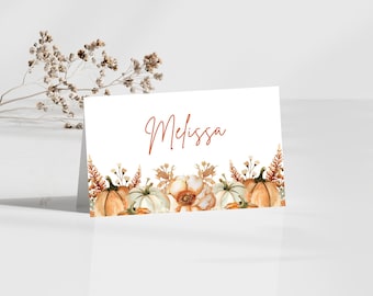 May include: A white place card with the name "Melissa" written in cursive in orange. The card is decorated with a border of watercolor pumpkins, flowers, and leaves.