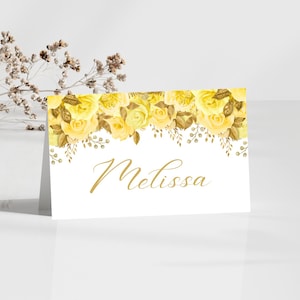 Pode incluir: Um cartão de lugar branco com um design floral de rosas amarelas e detalhes dourados. O nome "Melissa" está escrito em dourado. Um ramo de flores secas está no fundo.