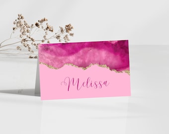 May include: A pink place card with a watercolor design in shades of pink and magenta, accented with gold glitter. The name "Melissa" is written in cursive script. The card is standing upright.
