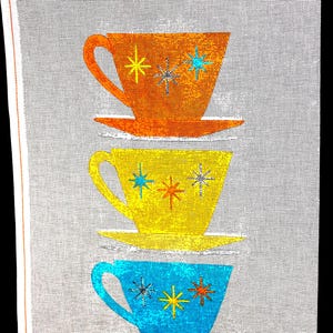 May include: A stack of three retro-style teacups in orange, yellow, and blue. Each cup features a starburst design in contrasting colors. The cups are on saucers, creating a mid-century modern aesthetic.