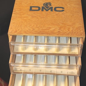 Puede incluir: Caja de almacenamiento de hilo de bordar DMC de madera con tres cajones de plástico transparente. La caja tiene el logotipo de DMC en la tapa y los cajones. Cada cajón contiene múltiples compartimentos.