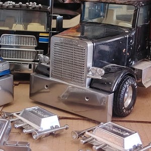 Puede incluir: Un camión de juguete negro con un parachoques delantero y una parrilla cromados. El camión tiene la palabra "Freightliner" impresa en la parte delantera. El camión está sentado sobre una superficie marrón con dos bocinas de juguete plateadas delante de él.