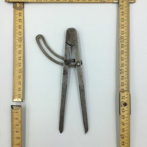 Puede incluir: Un compás de metal vintage con dos patas ajustables y un mango circular. El compás está hecho de metal y tiene un aspecto desgastado y envejecido.