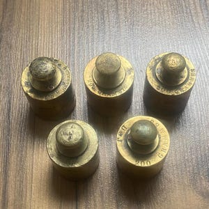 Può includere: Cinque pesi in ottone per una bilancia, ognuno con un peso diverso. I pesi sono rotondi e hanno una manopola sulla parte superiore. I pesi sono etichettati con il testo "200 GR", "200 GR", "200 GR", "200 GR" e "200 GR".
