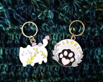 May include: Two enamel stitch markers with gold-colored rings. One is a white cat with purple mushrooms and yellow leaves. The other is a white circle with a black paw print, a pink heart, and a crescent moon design.