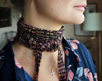 May include: A woman wearing a black and pink patterned crocheted neck scarf with a gold chain necklace and a small cross pendant.