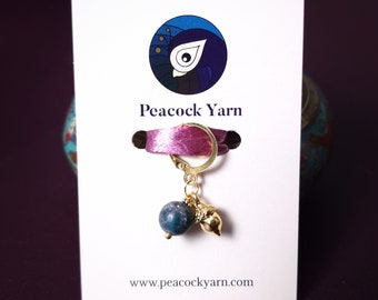 May include: A teal blue and gold stitch marker with an acorn charm. The stitch marker is attached to a pink ribbon and a gold ring. The card reads "Peacock Yarn" and "www.peacockyarn.com".