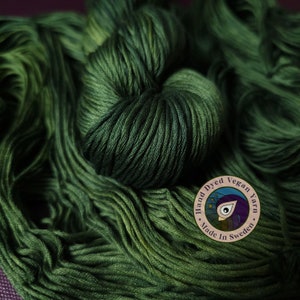 May include: A close-up of a skein of deep green hand-dyed vegan yarn. The yarn is made in Sweden and has a label that says "Hand Dyed Vegan Yarn Made in Sweden".