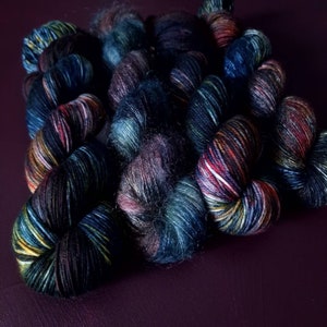 May include: A collection of hand-dyed yarn skeins in a variety of colors, including deep blues, purples, and reds. The yarn appears soft and textured, with subtle variations in color throughout each skein. The skeins are arranged on a dark purple surface.