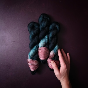 Op de afbeelding: Drie bollen garen in tinten van teal, groen en roze. Het garen is in een waaier vorm gerangschikt met de uiteinden van de bollen die elkaar overlappen.