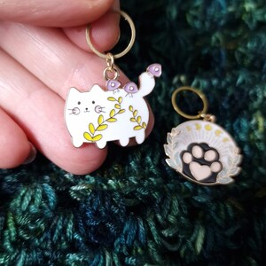 May include: Two enamel stitch markers. One is a white cat with yellow and green floral accents and purple mushrooms. The other is a paw print with a heart, surrounded by a wreath. Both have gold-coloured rings.