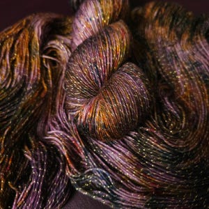 May include: A skein of yarn with a deep purple, brown, and gold color scheme. The yarn has a subtle shimmer and is made with a blend of fibers.