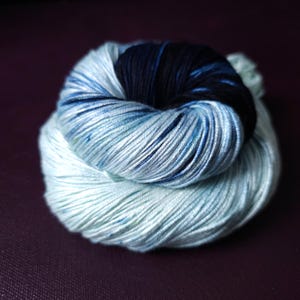 May include: A skein of hand-dyed yarn in shades of blue, teal, and navy. The yarn is tightly wound, showing a gradient effect from light to dark. The texture appears soft and smooth, suitable for knitting or crochet projects.