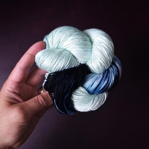 May include: A skein of yarn in shades of light blue, dark blue, and black. The yarn is tightly wound, creating a compact, textured bundle. The colors blend together, offering a gradient effect. The yarn is held in a hand against a dark background.