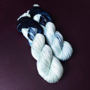 May include: Two skeins of yarn in a gradient of colors. The yarn transitions from a dark navy blue to a light blue-green. The yarn is twisted into a rope-like shape, with the ends of the yarn in a darker shade.
