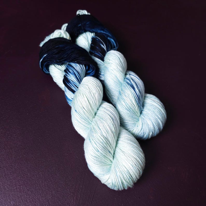 May include: Two skeins of yarn in a gradient of colors. The yarn transitions from a dark navy blue to a light blue-green. The yarn is twisted into a rope-like shape, with the ends of the yarn in a darker shade.