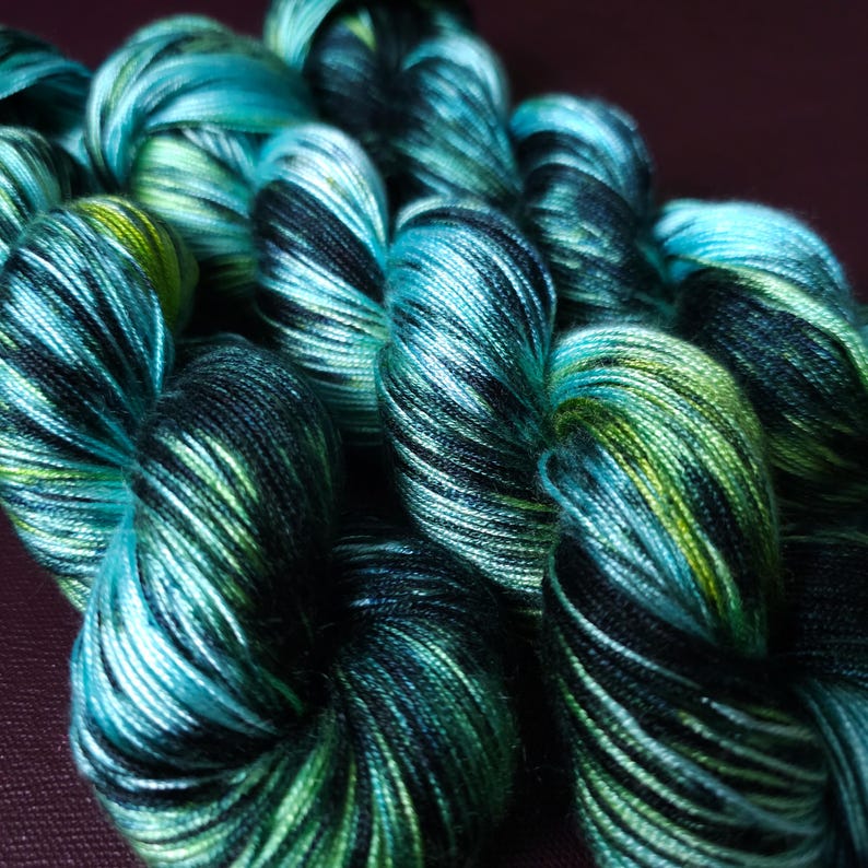 May include: Close-up of several skeins of yarn in shades of teal, black, and green. The yarn is tightly wound, with visible color variations creating a textured appearance. The color palette evokes a natural, aquatic theme.