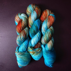 May include: Three skeins of yarn in shades of teal, blue, green, and orange. The yarn is twisted and piled on top of each other.