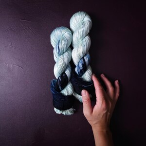May include: Two skeins of hand-dyed yarn in shades of light blue, navy, and black. The yarn is twisted into a rope-like shape, with the colors blending together. The yarn is on a dark purple background.