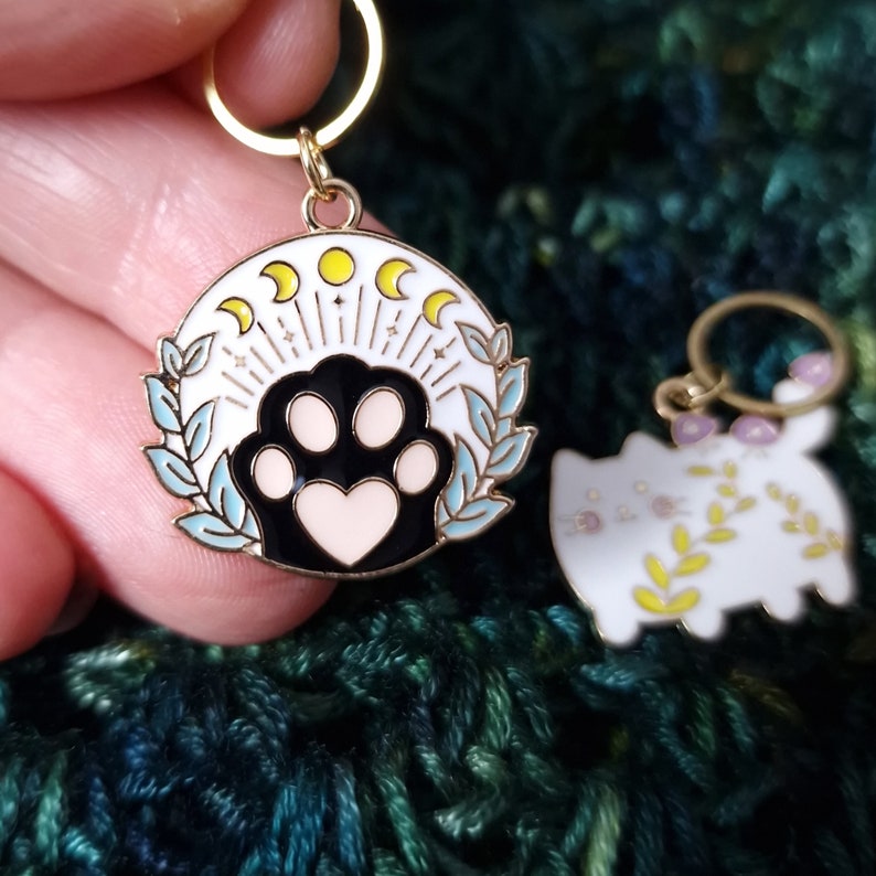 May include: Two gold-coloured stitch markers. One is a circular design with a black and white cat paw print with a heart, surrounded by a wreath and moon phases. The other is a white cat shape with floral accents.