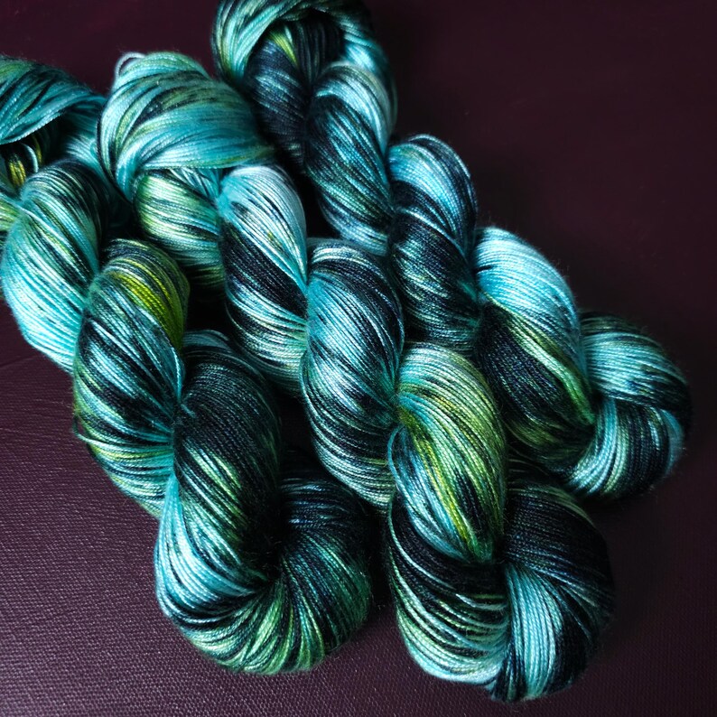 May include: Close-up of several skeins of hand-dyed yarn. The yarn is a mix of teal, black, and green, with a variegated pattern. The yarn is tightly wound and appears soft. The background is a dark purple.