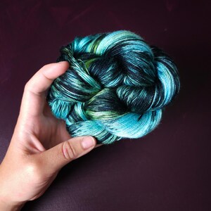 May include: A skein of hand-dyed yarn in shades of teal, turquoise, black, and green. The yarn is tightly wound and held in a hand, showcasing the color variations. This yarn is suitable for knitting, crocheting, and other fiber arts projects.