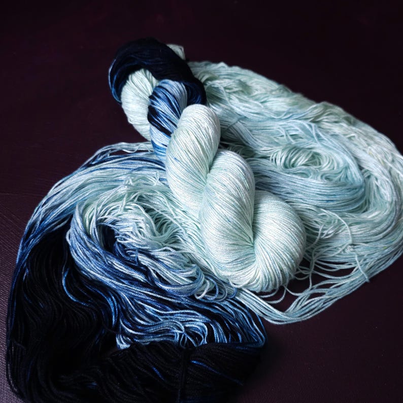May include: A close-up of several skeins of yarn in shades of blue and teal. The yarn is arranged in a loose, flowing pattern, with some skeins tightly wound. The color gradient ranges from deep navy to a light, airy teal.